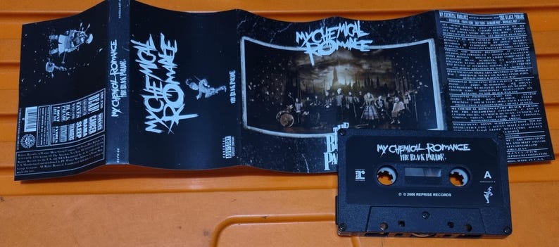Puede incluir: Una cinta de casete negra con el texto "My Chemical Romance The Black Parade" en la parte delantera. La cinta de casete est&aacute; en una caja de pl&aacute;stico con una imagen en blanco y negro de la banda en la parte delantera.