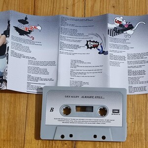 Może przedstawiać: Szara kaseta magnetofonowa z napisem "LILY ALLEN ALRIGHT, STILL" leży przed rozłożoną wkładką albumu. Wkładka zawiera zdjęcie kobiety i ilustracje z kresk&oacute;wek. Kaseta ma etykietę strony B.