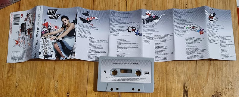 Może przedstawiać: Szara kaseta magnetofonowa z napisem "LILY ALLEN ALRIGHT, STILL" leży przed rozłożoną wkładką albumu. Wkładka zawiera zdjęcie kobiety i ilustracje z kresk&oacute;wek. Kaseta ma etykietę strony B.