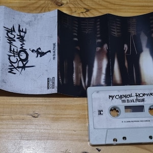 Puede incluir: Una cinta de casete con una etiqueta blanca y texto negro que dice "My Chemical Romance The Black Parade". La cinta de casete est&aacute; sobre una superficie de madera. La cinta de casete es parte de una imagen m&aacute;s grande que muestra una caja de cinta de casete con una imagen en blanco y negro de una banda en la portada.