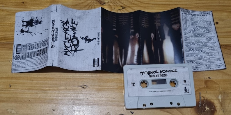 Puede incluir: Una cinta de casete con una etiqueta blanca y texto negro que dice "My Chemical Romance The Black Parade". La cinta de casete est&aacute; sobre una superficie de madera. La cinta de casete es parte de una imagen m&aacute;s grande que muestra una caja de cinta de casete con una imagen en blanco y negro de una banda en la portada.