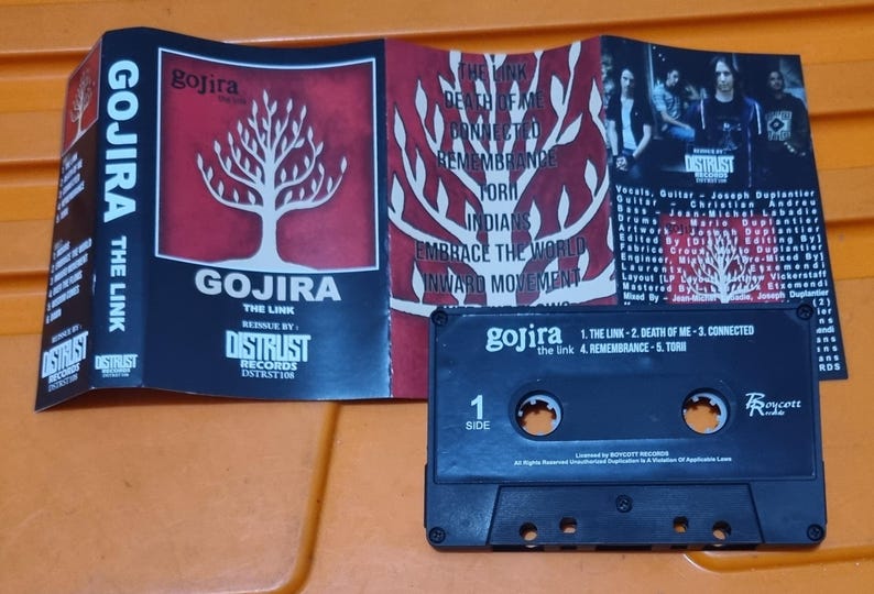 Pu&ograve; includere: Una cassetta audio nera con un'etichetta bianca. L'etichetta riporta il nome della band "Gojira" e il titolo dell'album "The Link". La cassetta &egrave; in una custodia di plastica con una finestra trasparente.