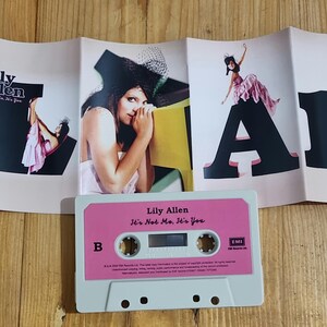 Może przedstawiać: Kaseta magnetofonowa i okładka albumu "It's Not Me, It's You" Lily Allen. Kaseta jest biała z r&oacute;żową etykietą. Okładka albumu przedstawia artystkę i duże czarne litery. Tło to drewniana powierzchnia.