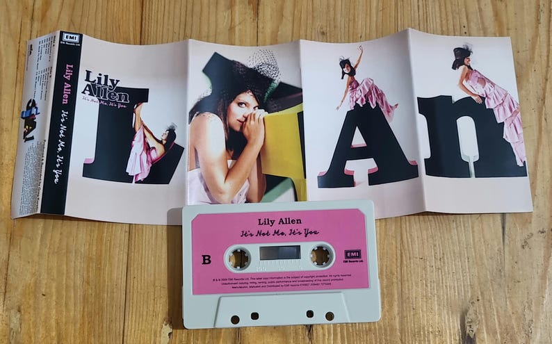 Może przedstawiać: Kaseta magnetofonowa i okładka albumu "It's Not Me, It's You" Lily Allen. Kaseta jest biała z r&oacute;żową etykietą. Okładka albumu przedstawia artystkę i duże czarne litery. Tło to drewniana powierzchnia.