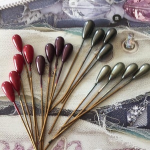 Peut inclure: Un ensemble de 17 épingles à chapeau dorées avec des têtes en verre en forme de larme rouge et verte. Les épingles sont disposées en éventail sur un fond de tissu blanc.