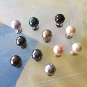 Puede incluir: Surtido de 11 botones de perla y piedra preciosa en varios colores, incluyendo negro, gris, blanco, marrón y beige. Los botones se muestran sobre una superficie de color amarillo claro y azul.