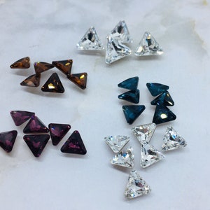 May include: Assortment of triangle-shaped rhinestones in various colours, including clear, purple, brown, and blue. The rhinestones are arranged in groups on a white surface.