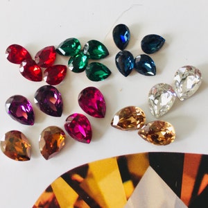 May include: Assortment of pear-shaped glass rhinestones in various colors, including red, green, blue, and clear. The rhinestones are arranged on a white surface.