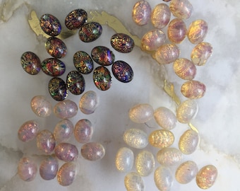 May include: A collection of oval-shaped cabochons arranged in four distinct groups. The beads feature a mix of dark blue with multicolored iridescent flecks, translucent white with pinkish hues, and pale yellow with shimmering gold-toned inclusions.