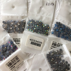 May include: Assortment of small clear plastic bags containing various colors of rhinestones. Each bag has a label with the text "SWAROVSKI" and a barcode. The bags are on a white surface.