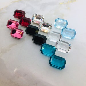 May include: Assortment of colorful emerald cut gemstones, including red, pink, black, blue, and clear. The gemstones are arranged in a triangular pattern on a white marble surface.