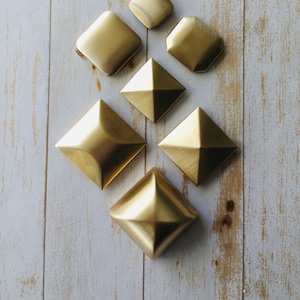 Op de afbeelding: Zeven goudkleurige metalen decoratieve stukken in verschillende geometrische vormen, waaronder vierkanten en piramides, gerangschikt op een lichte houten achtergrond.