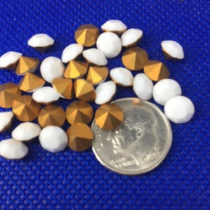 Könnte beinhalten: Eine Ansammlung goldener und weißer, facettierter und abgerundeter Perlen, verstreut auf einer blauen Oberfläche. Die goldenen Perlen haben ein metallisches Finish, während die weißen Perlen glatt sind. Eine Münze dient als Größenreferenz.