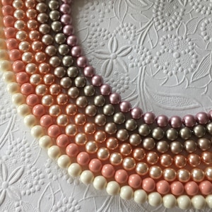May include: Multiple strands of round, smooth, lustrous beads arranged in a curved, layered pattern. The beads feature a gradient of colors including cream, soft peach, light pink, metallic bronze, and mauve, creating a multi-toned display.