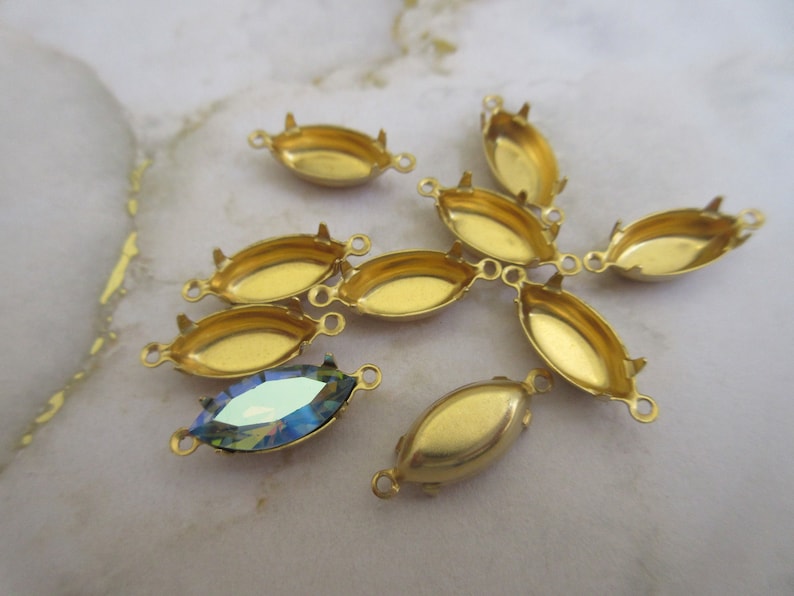 Puede incluir: Un grupo de ocho engastes de metal dorado para piedras en forma de marquesa. Los engastes tienen dos bucles a cada lado para sujetar a las joyas. Un engaste tiene una piedra en forma de marquesa azul iridiscente.