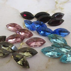 Peut inclure: Assortiment de strass en verre de forme marquise de différentes couleurs, bleu, rose, marron et bordeaux. Les strass sont disposés sur une surface blanche.