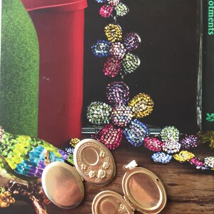 May include: Two gold oval locket pendants with intricate floral designs. One locket is open, revealing a blank interior. The lockets are on a wooden surface.