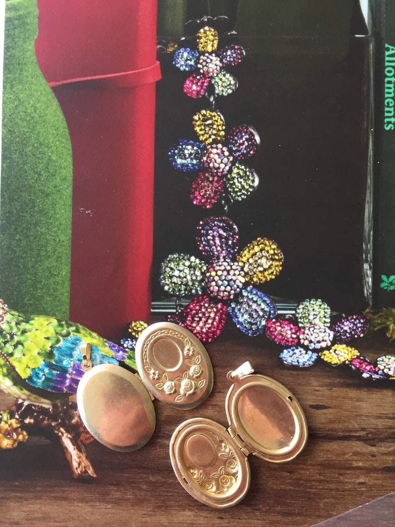 May include: Two gold oval locket pendants with intricate floral designs. One locket is open, revealing a blank interior. The lockets are on a wooden surface.