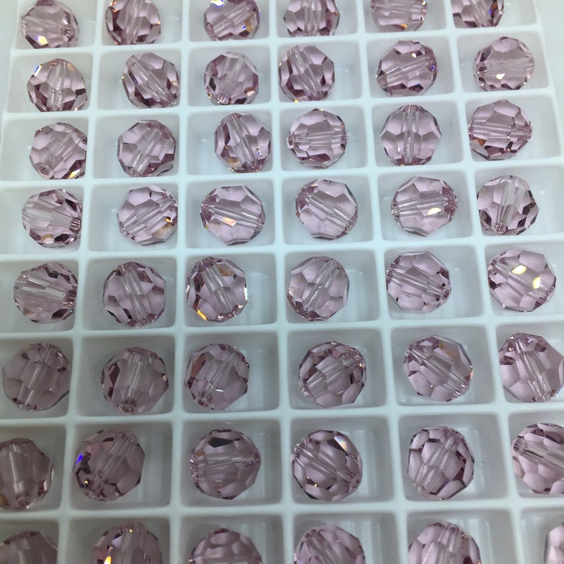 May include: A close-up view of a container filled with small, round, light pink glass beads. The beads are arranged in a grid pattern and have a faceted design.
