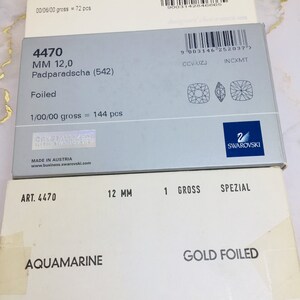 May include: Two boxes of Swarovski crystals. The top box is white with the text "4470 12 mm Violet (371) Foiled 00/06/00 gross = 72 pcs" and a barcode. The bottom box is white with the text "ART. 4470 12 MM 1 GROSS SPEZIAL AQUAMARINE GOLD FOILED SWAROVSKI Made in Austria".