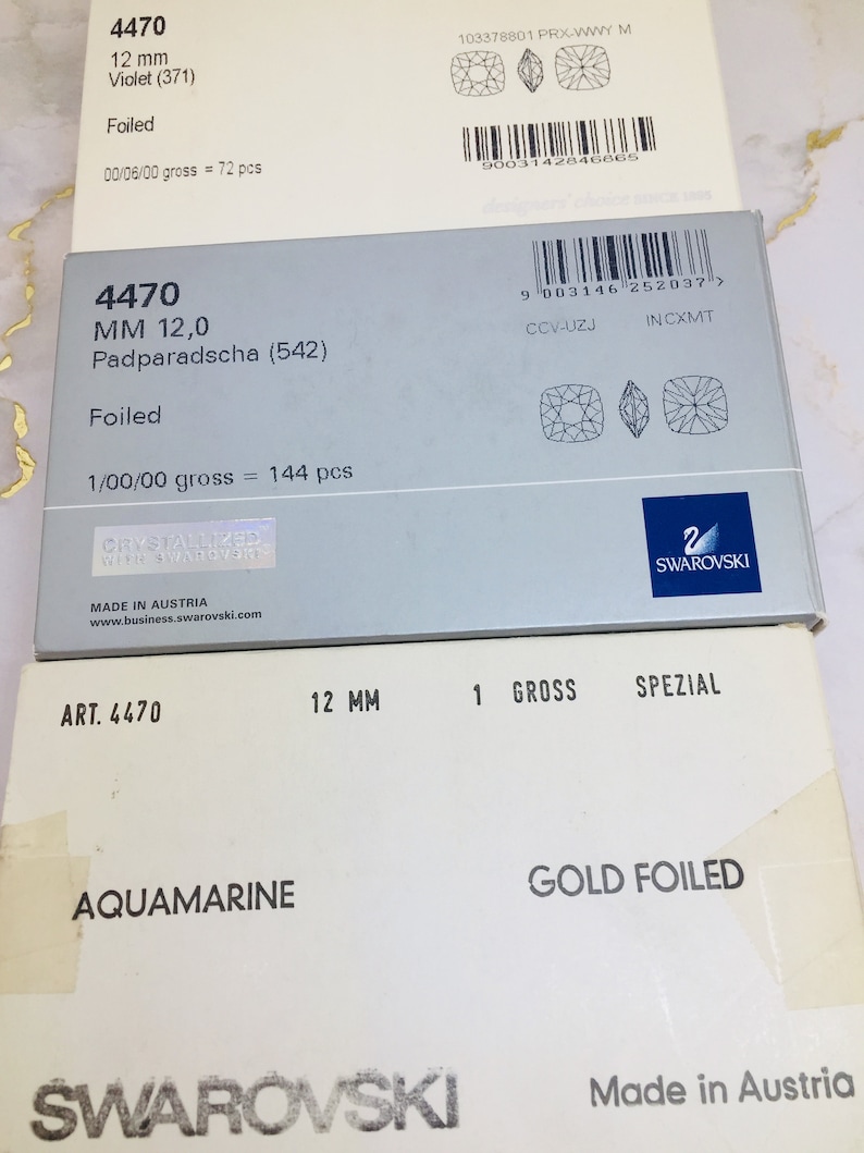 May include: Two boxes of Swarovski crystals. The top box is white with the text "4470 12 mm Violet (371) Foiled 00/06/00 gross = 72 pcs" and a barcode. The bottom box is white with the text "ART. 4470 12 MM 1 GROSS SPEZIAL AQUAMARINE GOLD FOILED SWAROVSKI Made in Austria".