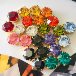 May include: A collection of round, faceted, multi-colored glass stones set in gold-toned metal settings. The assortment includes red, yellow, orange, green, blue, purple, pink, black, and clear stones arranged in a cluster on a white surface.