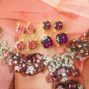 May include: An assortment of sparkling jewelry components in shades of pink and purple. The components include square and oval-shaped crystals with gold-colored metal settings. The image also features a section of a larger, ornate necklace with similar crystals.