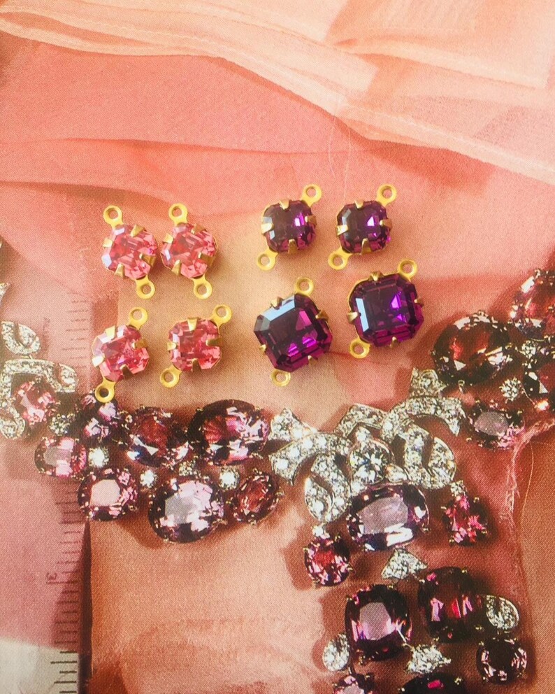 May include: An assortment of sparkling jewelry components in shades of pink and purple. The components include square and oval-shaped crystals with gold-colored metal settings. The image also features a section of a larger, ornate necklace with similar crystals.