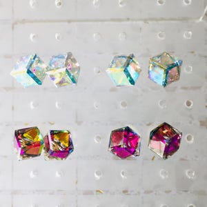 May include: Six iridescent cube-shaped earrings displayed on a white background. Two pairs are clear with rainbow iridescent colors, and one pair is a deep purple with rainbow iridescent colors.