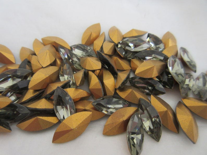 Puede incluir: Un mont&oacute;n de piedras de imitaci&oacute;n de forma de marquesa doradas y gris oscuro. Las piedras de imitaci&oacute;n son facetadas y tienen un acabado brillante.