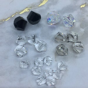 Może przedstawiać: Zestaw przezroczystych szklanych koralików o różnych szlifach i fasetach, w tym dwa czarne fasetowane koraliki.