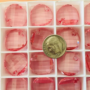 May include: A close-up of a box of pink round crystal beads with a US quarter for scale. The beads have a hole in the top for stringing.
