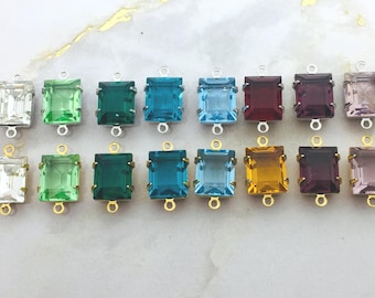 May include: A collection of rectangular gemstone connectors in various colors, including clear, green, teal, blue, red, purple, and yellow. Each connector has a gold-colored bail at the top and bottom for attaching to other components.