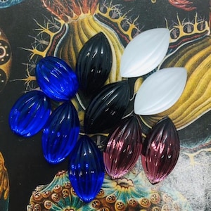 May include: A collection of ten marquise-shaped cabochons featuring a vertical ribbed texture. The set includes three translucent blue pieces, two opaque black pieces, two matte white pieces, and two translucent pink pieces with a metallic finish.