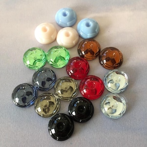 Könnte beinhalten: Sortiment von 16 runden Glasknöpfen in verschiedenen Farben, darunter Grün, Rot, Blau, Braun und Schwarz. Die Knöpfe haben eine erhabene Mitte und eine strukturierte Oberfläche.