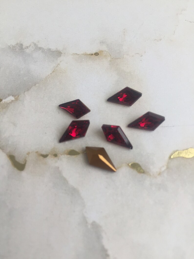 May include: Assortment of red and gold diamond-shaped rhinestones on a white marble background.