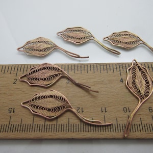 May include: Six gold-toned wire leaf-shaped charms, each measuring approximately 2 inches in length, are arranged on a wooden ruler.