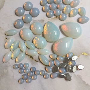May include: A collection of iridescent opal cabochons in various shapes and sizes, including round, oval, and teardrop. The opals are a milky white color with a hint of pink and blue.