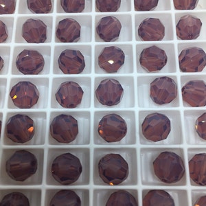May include: A close-up view of a container filled with round, faceted, dark purple glass beads. The beads are arranged in a grid pattern.
