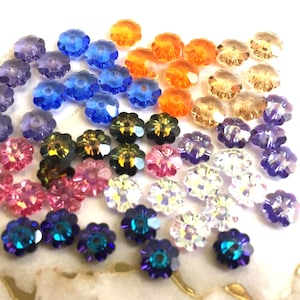 May include: A collection of small, faceted flower-shaped beads in a variety of colors including blue, purple, orange, pink, clear, and iridescent tones. Each bead features a central hole and a multi-petaled design with reflective surfaces.