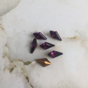 May include: A group of six purple and gold diamond-shaped rhinestones on a white marble surface.