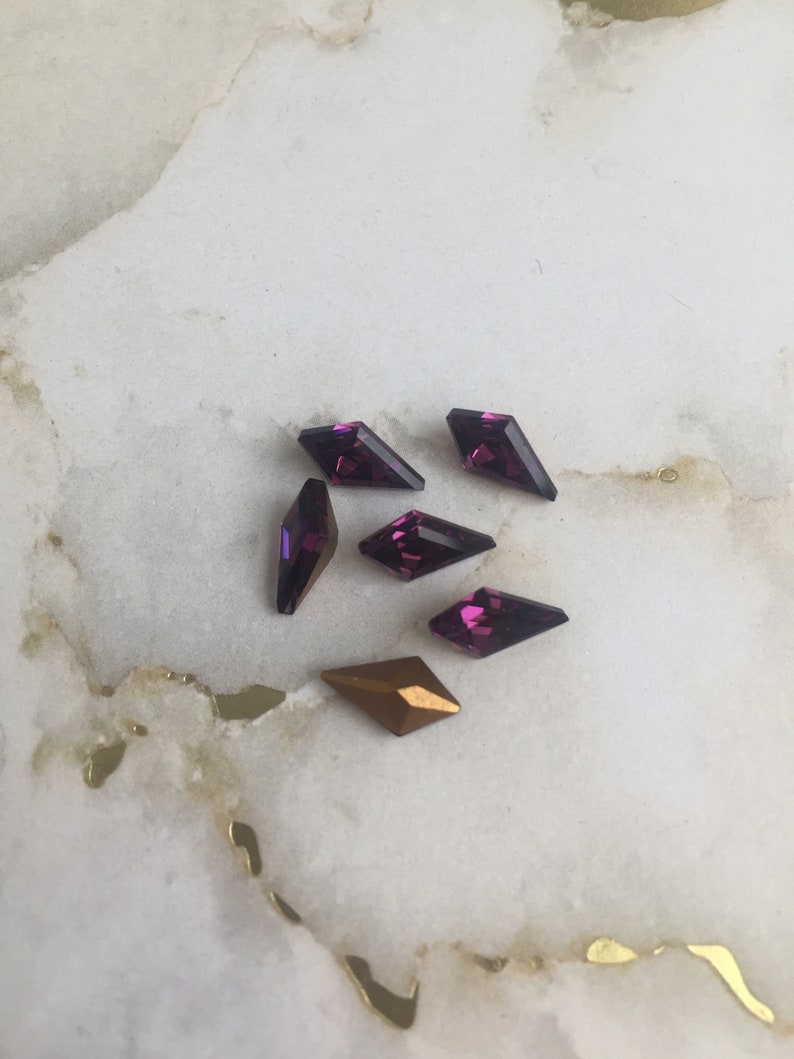 May include: A group of six purple and gold diamond-shaped rhinestones on a white marble surface.