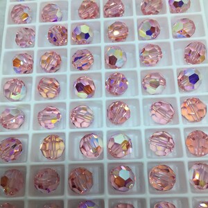 Peut inclure: Gros plan sur un plateau de perles rondes en verre rose. Les perles sont dispos&eacute;es en grille et ont une finition facet&eacute;e et iris&eacute;e.