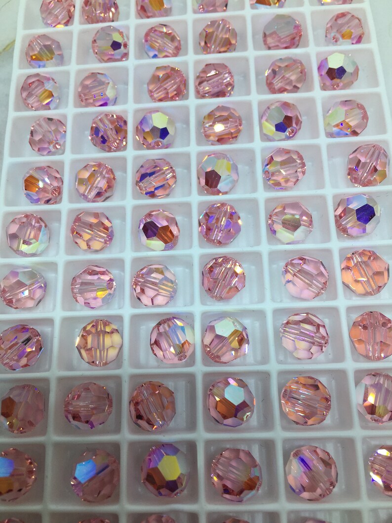 Peut inclure: Gros plan sur un plateau de perles rondes en verre rose. Les perles sont dispos&eacute;es en grille et ont une finition facet&eacute;e et iris&eacute;e.