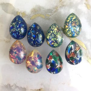 Puede incluir: Una colección de ocho cabujones en forma de lágrima con un acabado liso y brillante. Cada pieza contiene escamas de lámina metálica incrustadas en varios colores, incluyendo azul profundo, verde esmeralda, oro iridiscente y tonos multicolores.