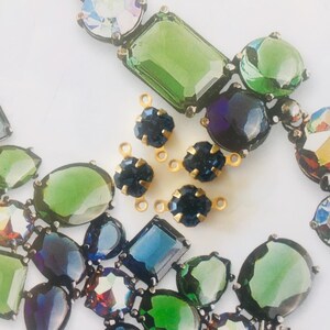 May include: Close-up of a necklace and loose jewelry components. The necklace features large, colorful, faceted gemstones in shades of green, blue, and iridescent hues. Several small, dark blue, flower-shaped gems with gold settings are also present.
