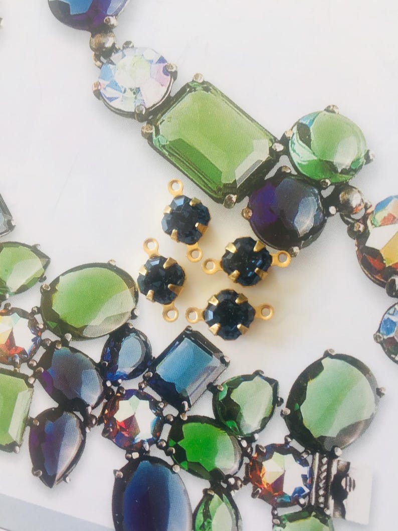 May include: Close-up of a necklace and loose jewelry components. The necklace features large, colorful, faceted gemstones in shades of green, blue, and iridescent hues. Several small, dark blue, flower-shaped gems with gold settings are also present.