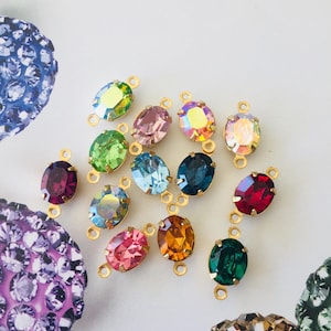 May include: A selection of oval-shaped, colourful crystal connectors with gold-coloured loops. The crystals are in various colours, including green, pink, blue, and red. The crystals are arranged on a white background.