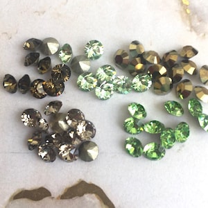 Op de afbeelding: Een verzameling ronde, gefacetteerde kristallen gerangschikt in vier afzonderlijke groepen. De stenen hebben verschillende kleuren, waaronder tinten donkerbruin, metallic brons, licht muntgroen en levendig limoengroen, allemaal met reflecterende oppervlakken.