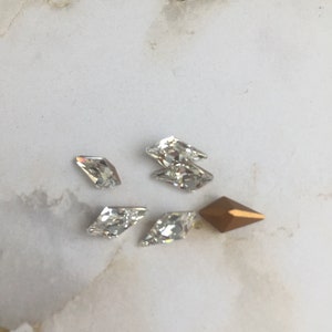 May include: Assortment of clear and gold rhinestones in various shapes, including a diamond shape, a teardrop shape, and a rectangular shape.